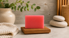 A rectangular pink soap bar displayed on a small wooden soap dish, styled on a tabletop with a ceramic vase, green leaves, stacked spa stones, and a folded knit towel in the background.